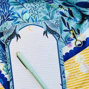 Puede incluir: Un bloc de notas rayado con un diseño de ballena y luna, un bolígrafo verde claro y una llave decorativa descansan sobre una superficie estampada azul y amarilla. El bloc de notas tiene un borde azul con ilustraciones de ballenas y una luna.