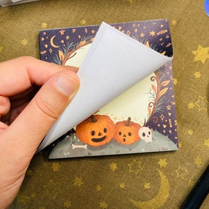 May include: A square notepad with a Halloween-themed illustration. The illustration features two orange pumpkins with carved faces, a skull, and a bone on a grassy hill. The background is a dark blue with stars, moons, and decorative elements.