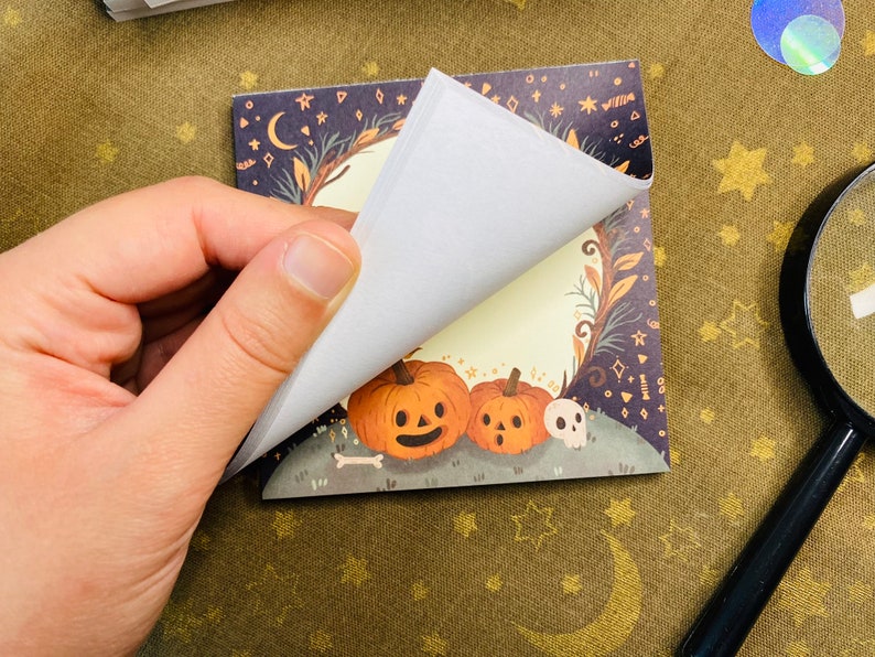 May include: A square notepad with a Halloween-themed illustration. The illustration features two orange pumpkins with carved faces, a skull, and a bone on a grassy hill. The background is a dark blue with stars, moons, and decorative elements.