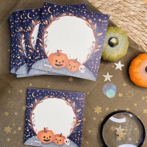 May include: A stack of Halloween-themed cards with a dark blue border featuring moons, stars, and leaves. The cards have a circular blank space with pumpkins and a skull at the bottom. Decorative pumpkins and stars are also present.