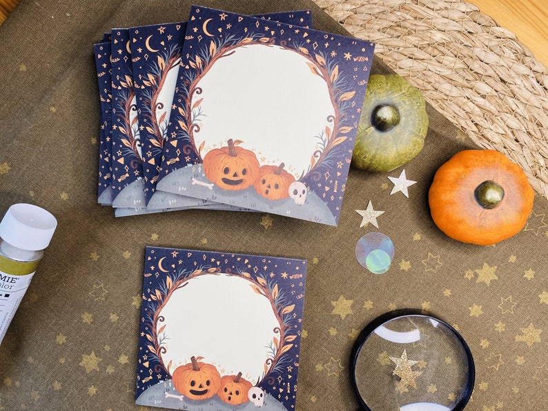 May include: A stack of Halloween-themed cards with a dark blue border featuring moons, stars, and leaves. The cards have a circular blank space with pumpkins and a skull at the bottom. Decorative pumpkins and stars are also present.
