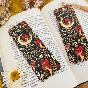 May include: Two black bookmark with a crescent moon and mushrooms on a dark background with gold accents. The bookmarks have a tassel on the top.