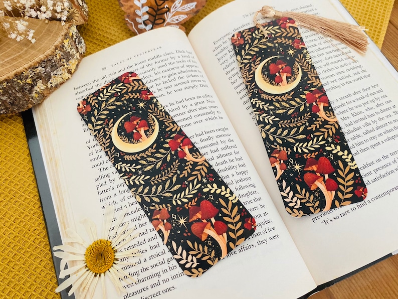 May include: Two black bookmark with a crescent moon and mushrooms on a dark background with gold accents. The bookmarks have a tassel on the top.