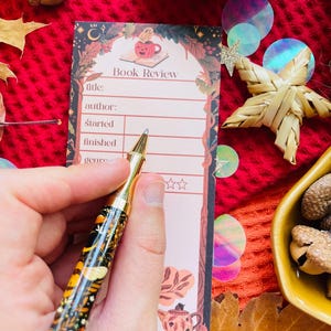 K&ouml;nnte beinhalten: Ein dekorativer Stift mit goldener Spitze und schwarzem Schaft mit floralem und Insektenmuster liegt &uuml;ber einem "Book Review" Notizblock. Der Notizblock hat eine K&uuml;rbisillustration und Platz zum Ausf&uuml;llen.