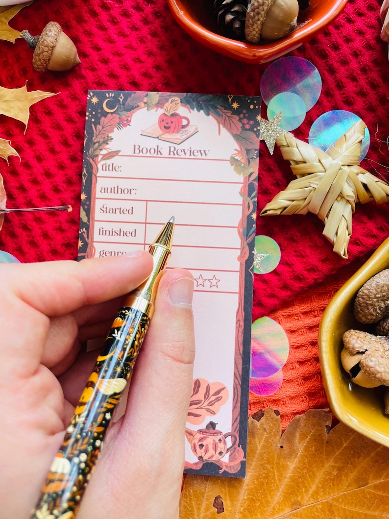 K&ouml;nnte beinhalten: Ein dekorativer Stift mit goldener Spitze und schwarzem Schaft mit floralem und Insektenmuster liegt &uuml;ber einem "Book Review" Notizblock. Der Notizblock hat eine K&uuml;rbisillustration und Platz zum Ausf&uuml;llen.