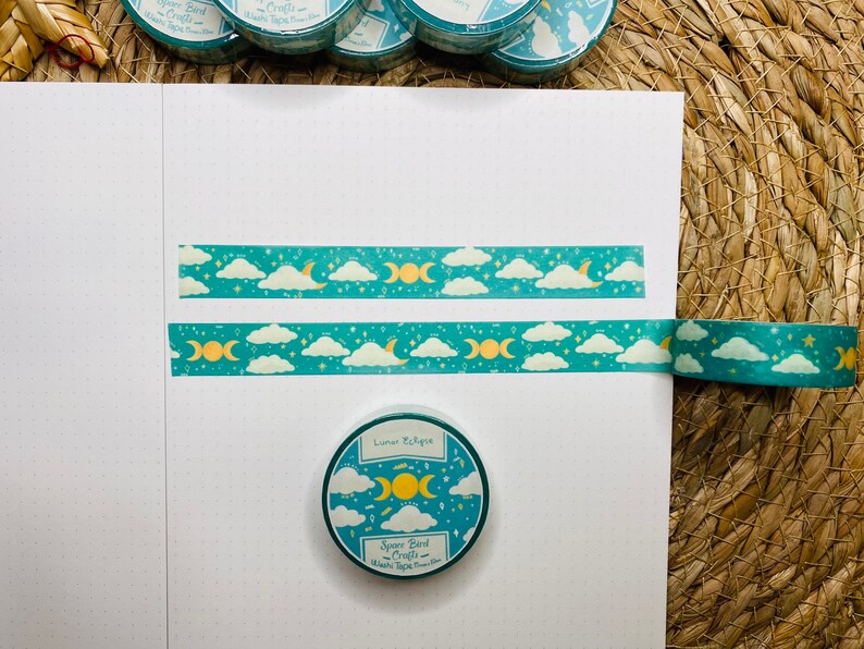 May include: A roll of teal washi tape with a moon and cloud pattern. The tape is unrolled and shows the design. The tape is labeled "Lunar Eclipse" and "Space Bird Crafts".