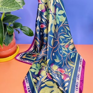 Può includere: Una sciarpa quadrata di seta con un motivo di fichi e foglie nei toni del blu, verde, viola e rosa. La sciarpa ha uno sfondo blu scuro con un bordo e viene tenuta in aria.