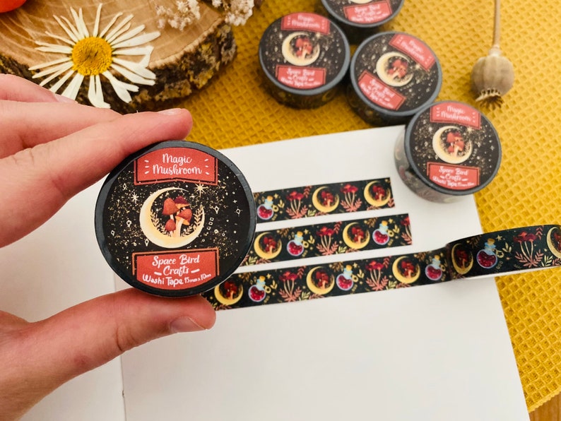 Op de afbeelding: Een rol zwarte en rode washi tape met een ontwerp van paddenstoelen, manen en sterren. De tape is gelabeld "Magic Mushroom - Space Bird Crafts - Washi Tape (10mm)"