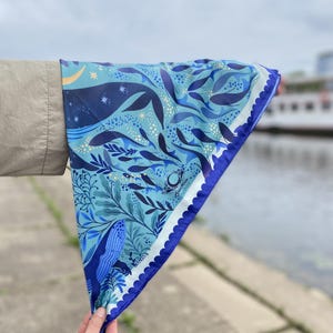Puede incluir: Un pañuelo azul y blanco con un diseño de ballenas y plantas acuáticas. El pañuelo tiene un borde festoneado y un borde azul oscuro. El diseño incluye ballenas, peces y estrellas, creando un tema náutico.
