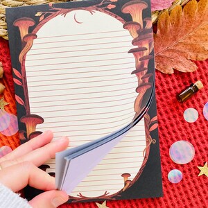 May include: A lined notepad with a mushroom and crescent moon design. The notepad has a black border with brown and orange mushrooms. The pages are cream-colored with horizontal lines. The notepad is being held open, revealing the blank pages.