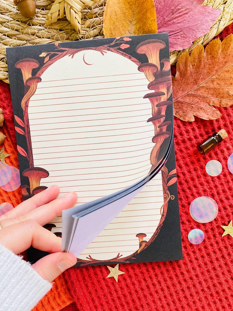 May include: A lined notepad with a mushroom and crescent moon design. The notepad has a black border with brown and orange mushrooms. The pages are cream-colored with horizontal lines. The notepad is being held open, revealing the blank pages.