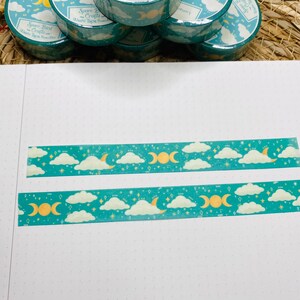 May include: Two rolls of teal washi tape with a gold moon and cloud pattern. The tape is on a white surface with a dotted pattern.