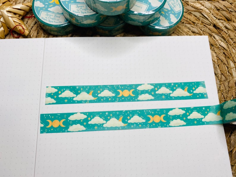 May include: Two rolls of teal washi tape with a gold moon and cloud pattern. The tape is on a white surface with a dotted pattern.