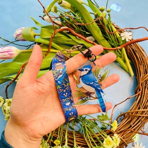 Può includere: Un portachiavi con un disegno di ghiandaia azzurra blu e bianca, attaccato a un cordino blu con un motivo celeste. Il portachiavi è tenuto davanti a una ghirlanda floreale con sfondo blu.