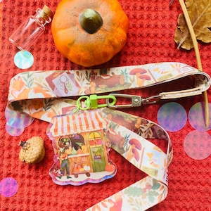 Puede incluir: Un cordón blanco y naranja con estampado y un cierre verde y un dije de acrílico transparente que representa una pequeña tienda con un toldo a rayas y una calabaza. El dije está unido al cordón con un anillo plateado.