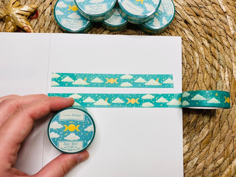May include: A roll of teal washi tape with a moon and cloud design. The tape is labeled "Lunar Eclipse" and "Space Bird Crafts".