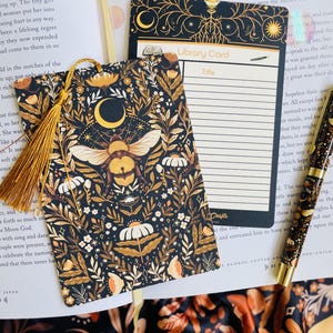 May include: A black bookmark with a gold tassel, featuring a bee and floral design. A matching library card and pen are also visible. The bookmark and card have a celestial theme with moons and stars.