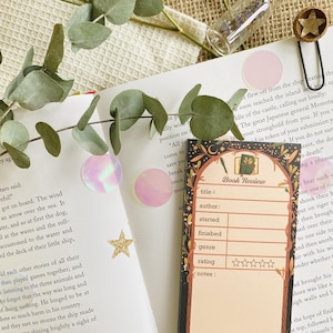 May include: A book review bookmark with a decorative border, featuring fields for title, author, and rating. The bookmark is placed on an open book with eucalyptus leaves, pink circles, and star-shaped confetti.