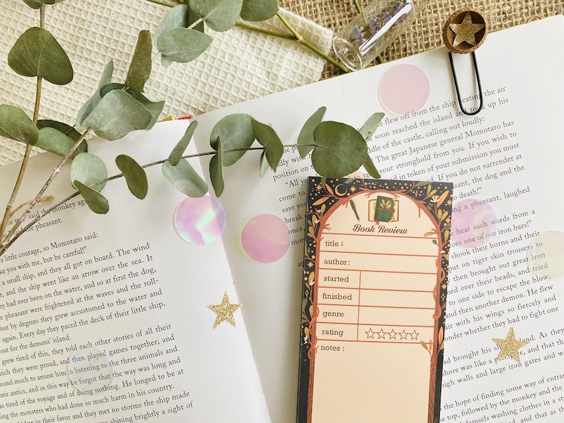 May include: A book review bookmark with a decorative border, featuring fields for title, author, and rating. The bookmark is placed on an open book with eucalyptus leaves, pink circles, and star-shaped confetti.