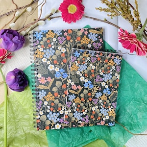 Puede incluir: Dos cuadernos de espiral con tapas negras y un patrón floral. Los cuadernos están sobre un fondo verde y blanco con flores rosas.