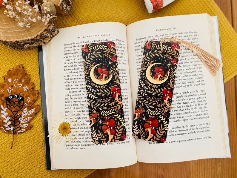 May include: Two black and gold bookmarks with a crescent moon and mushrooms on a dark background. The bookmarks have a tassel.