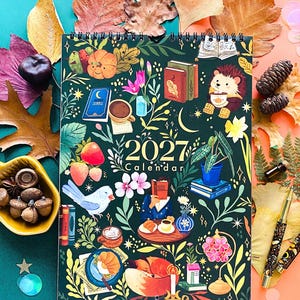 Puede incluir: Un calendario de pared 2027 con fondo verde oscuro que presenta ilustraciones coloridas de un erizo, un zorro, libros, flores, pájaros y alimentos. El calendario está encuadernado en espiral en la parte superior y muestra el año 2027 en letras doradas.