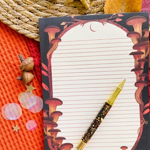 May include: A lined notepad with a mushroom illustration and a gold-accented pen with a floral design. The notepad has a black border with red and brown mushrooms. The pen is black with gold accents and a floral pattern.