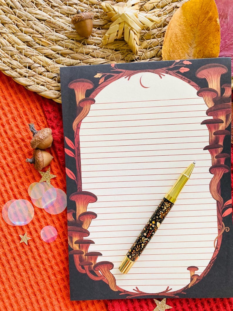 May include: A lined notepad with a mushroom illustration and a gold-accented pen with a floral design. The notepad has a black border with red and brown mushrooms. The pen is black with gold accents and a floral pattern.