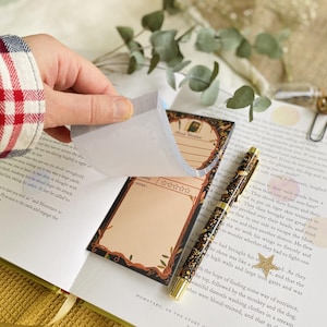 May include: A hand lifts a page from a notepad with a decorative border and space for notes. A patterned pen and open book with text are also visible. The notepad has a dark border with a light brown interior.