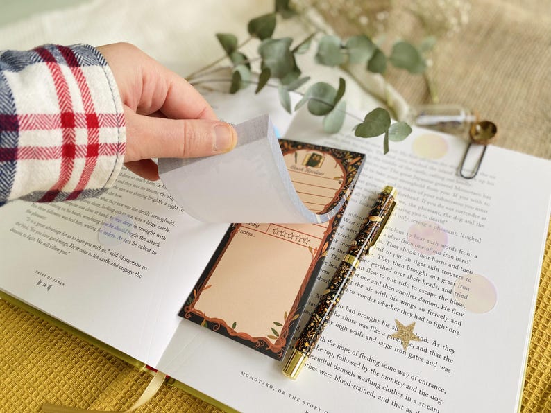 May include: A hand lifts a page from a notepad with a decorative border and space for notes. A patterned pen and open book with text are also visible. The notepad has a dark border with a light brown interior.