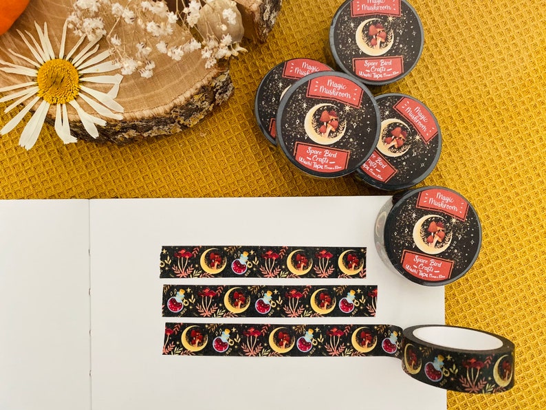Op de afbeelding: Drie rollen zwarte en gouden washi tape met een herhalend patroon van rode paddenstoelen, halve manen en sterren. De tape is gelabeld met "Magic Mushroom" en "Space Bird Crafts".