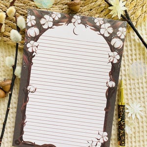 Pu&ograve; includere: Un blocco note a righe con un bordo decorativo di viti marroni e fiori di cotone bianchi. Una penna dorata e nera con disegni floreali &egrave; appoggiata accanto al blocco note. Il blocco note ha una parte superiore curva e un motivo a mezzaluna.