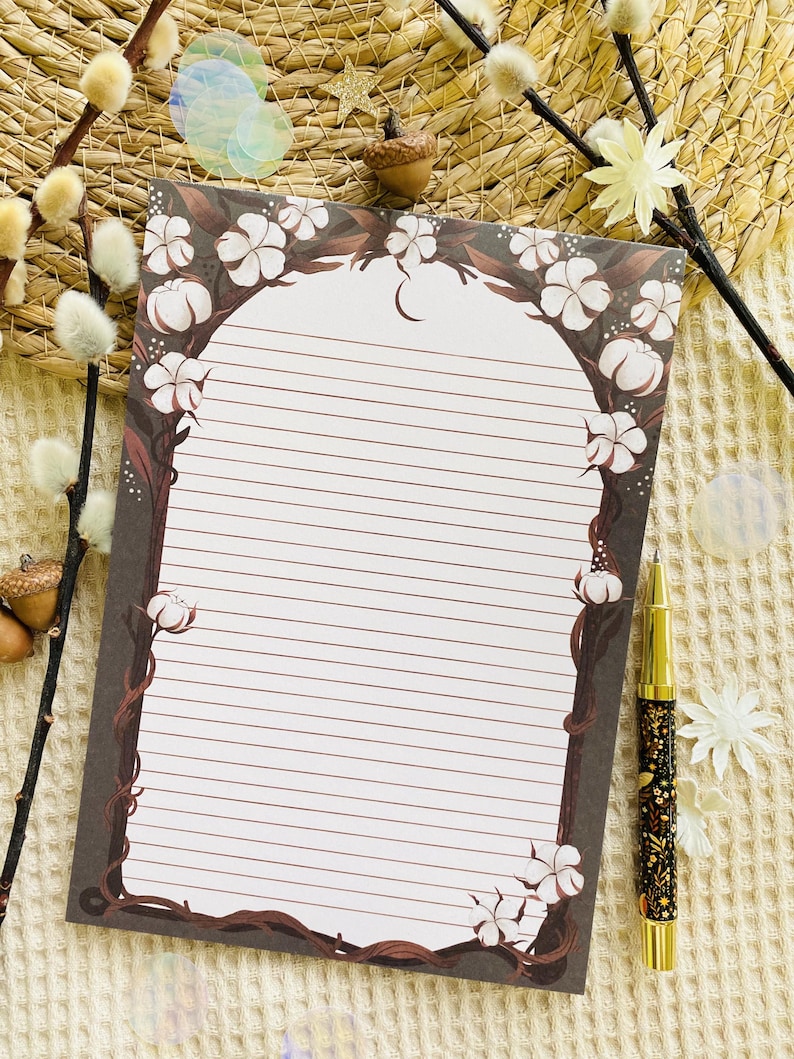 Pu&ograve; includere: Un blocco note a righe con un bordo decorativo di viti marroni e fiori di cotone bianchi. Una penna dorata e nera con disegni floreali &egrave; appoggiata accanto al blocco note. Il blocco note ha una parte superiore curva e un motivo a mezzaluna.