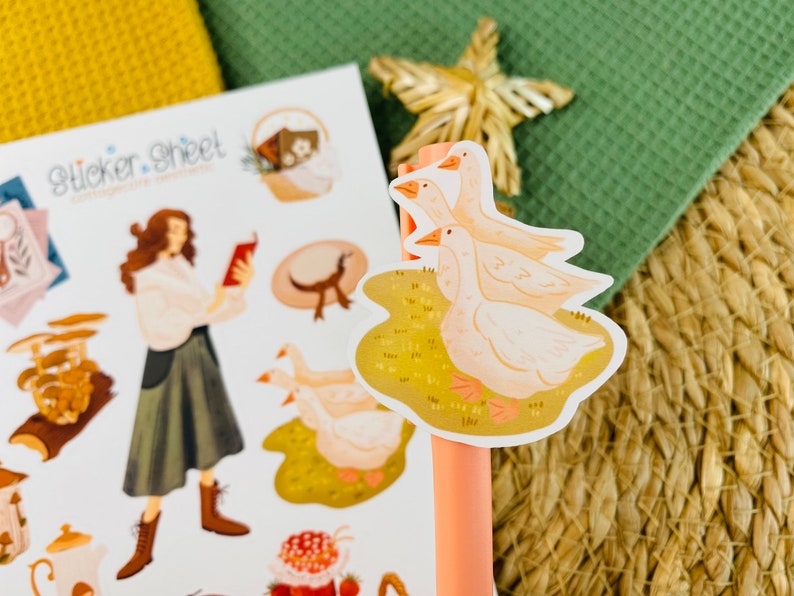 以下が含まれることがあります： 黄色い草のフィールドに3羽の白いガチョウが描かれた、幻想的なイラストのステッカーシート。ステッカーシートには、本を読んでいる女性、キノコの群れ、ティーポット、帽子も描かれています。