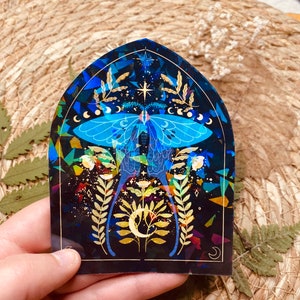 K&ouml;nnte beinhalten: Ein holografischer Aufkleber mit einer blauen und goldenen Motte mit einem Halbmond und Sternen auf schwarzem Hintergrund.