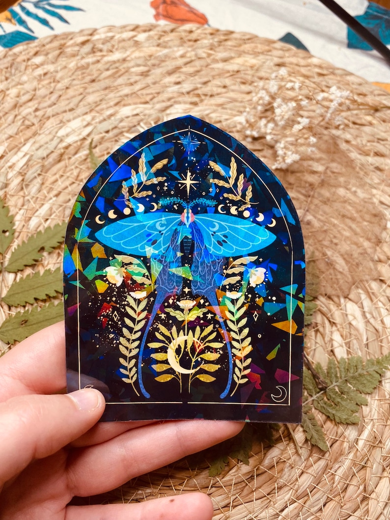 K&ouml;nnte beinhalten: Ein holografischer Aufkleber mit einer blauen und goldenen Motte mit einem Halbmond und Sternen auf schwarzem Hintergrund.