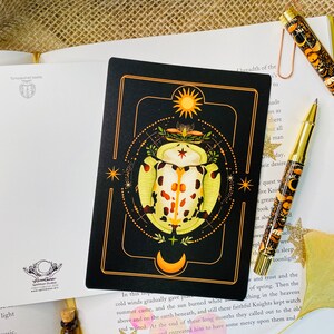 May include: A black card with a gold border features a stylized illustration of a beetle with orange and white markings. The beetle is surrounded by a circle of stars and a crescent moon. The card has the text "In this top" at the bottom.