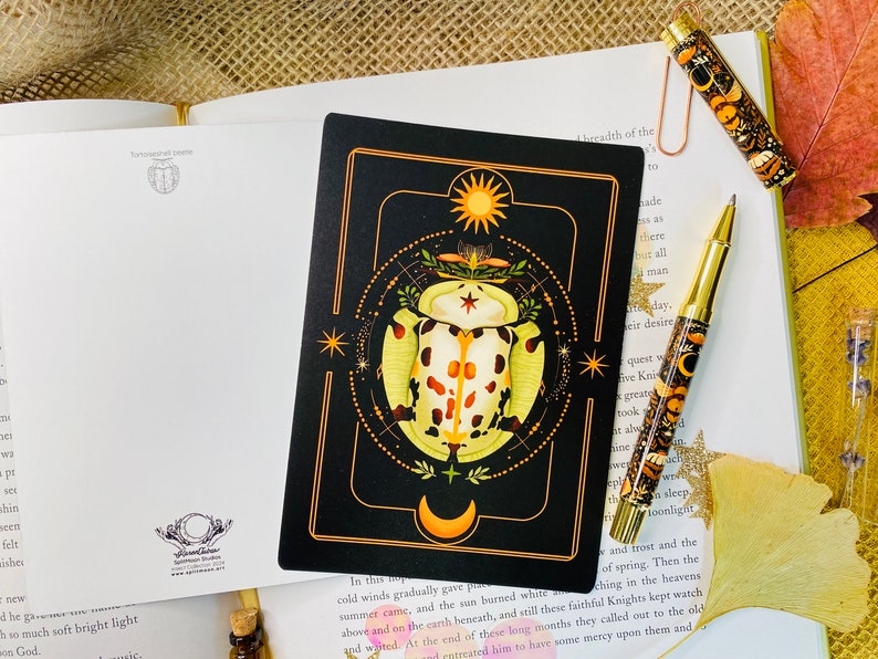 May include: A black card with a gold border features a stylized illustration of a beetle with orange and white markings. The beetle is surrounded by a circle of stars and a crescent moon. The card has the text "In this top" at the bottom.