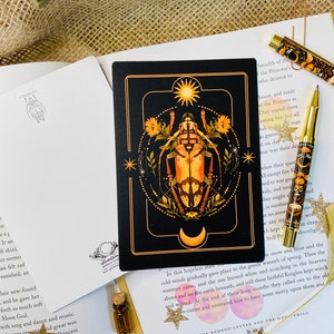 May include: A black card with a gold frame features a detailed illustration of a beetle with a sun and moon above it. The beetle is surrounded by flowers and stars. The card has a black background with gold accents.