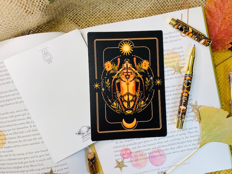 May include: A black card with a gold frame features a detailed illustration of a beetle with a sun and moon above it. The beetle is surrounded by flowers and stars. The card has a black background with gold accents.