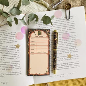 May include: A book review notepad with a decorative border, a matching pen, and a star-shaped paper clip rest on an open book. The notepad has spaces for title, author, and rating. The pen is dark with gold accents.