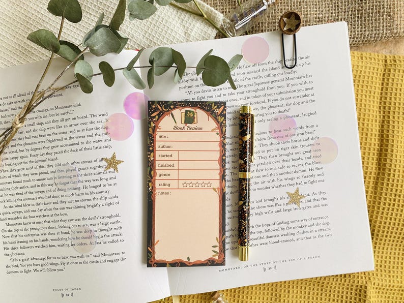 May include: A book review notepad with a decorative border, a matching pen, and a star-shaped paper clip rest on an open book. The notepad has spaces for title, author, and rating. The pen is dark with gold accents.