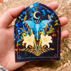 K&ouml;nnte beinhalten: Ein Aufkleber mit holografischem Finish, der einen blau-wei&szlig;en Nachtfalter mit ausgebreiteten Fl&uuml;geln zeigt, umgeben von gelben Blumen und einem Halbmond auf schwarzem Hintergrund.