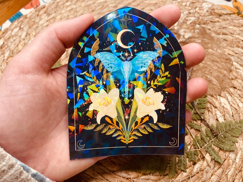 K&ouml;nnte beinhalten: Ein Aufkleber mit holografischem Finish, der einen blau-wei&szlig;en Nachtfalter mit ausgebreiteten Fl&uuml;geln zeigt, umgeben von gelben Blumen und einem Halbmond auf schwarzem Hintergrund.