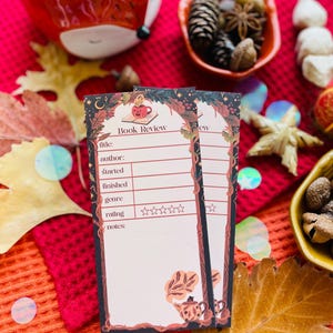 K&ouml;nnte beinhalten: Zwei rechteckige Buchbesprechungskarten mit einem dekorativen Rand mit einer Teetasse und floralem Design. Die Karten haben Platz f&uuml;r Titel, Autor und Bewertung. Der Hintergrund umfasst Herbstlaub und dekorative Schalen.