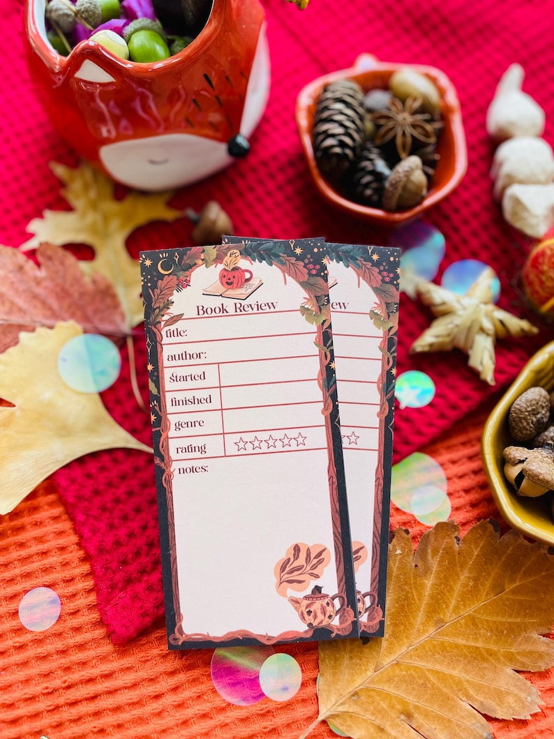 K&ouml;nnte beinhalten: Zwei rechteckige Buchbesprechungskarten mit einem dekorativen Rand mit einer Teetasse und floralem Design. Die Karten haben Platz f&uuml;r Titel, Autor und Bewertung. Der Hintergrund umfasst Herbstlaub und dekorative Schalen.