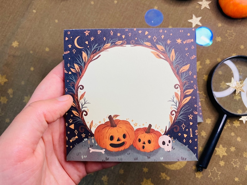 May include: Square Halloween-themed notepad featuring a pumpkin illustration. The design includes two orange pumpkins with carved faces, a skull, and a bone on a gray hill, surrounded by a floral wreath and a dark blue background with stars.
