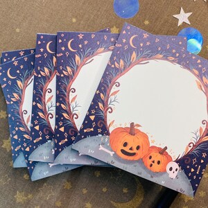 May include: A stack of Halloween-themed cards with a dark blue background featuring moons, stars, and decorative elements. Each card has a circular frame with pumpkins, a skull, and a bone at the bottom. The cards are perfect for Halloween.