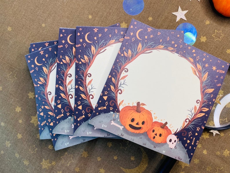 May include: A stack of Halloween-themed cards with a dark blue background featuring moons, stars, and decorative elements. Each card has a circular frame with pumpkins, a skull, and a bone at the bottom. The cards are perfect for Halloween.