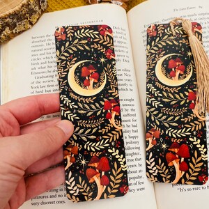 May include: A black bookmark with a gold crescent moon and mushrooms. The bookmark has a gold tassel.
